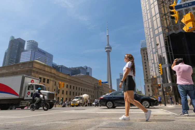 Mulher atravessando rua no centro de Toronto com a CN Tower ao fundo em um dia ensolarado.