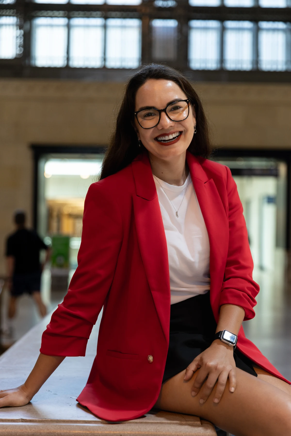 Débora Marques sorrindo, de óculos e blazer vermelho, sentada em um ambiente iluminado por grandes janelas.