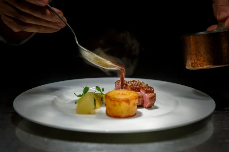 Prato refinado sendo finalizado por um chef com molho sobre carne e acompanhamento.