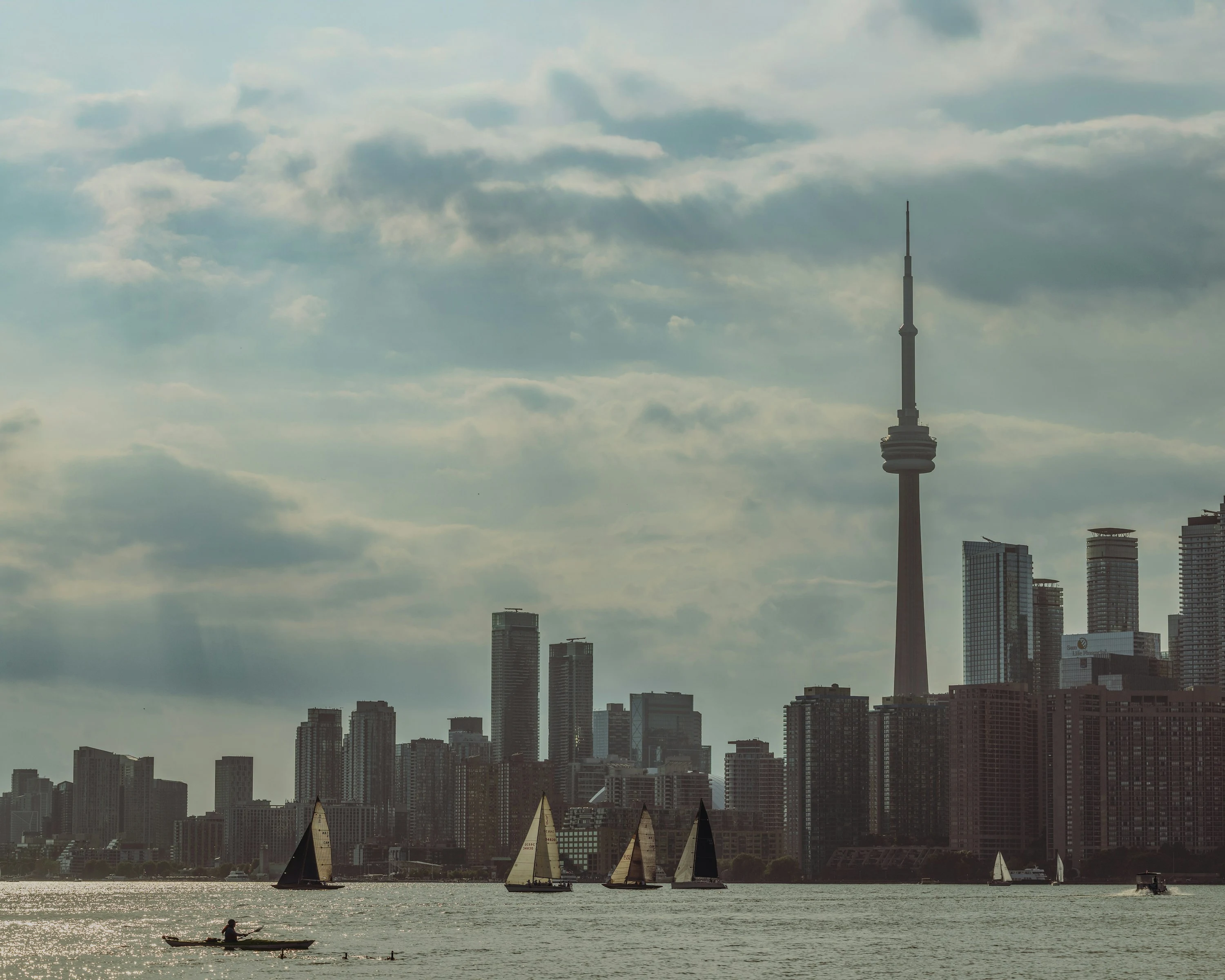 Veleiros e um caiaque no Lago Ontário, diante do skyline de Toronto sob um céu de nuvens.