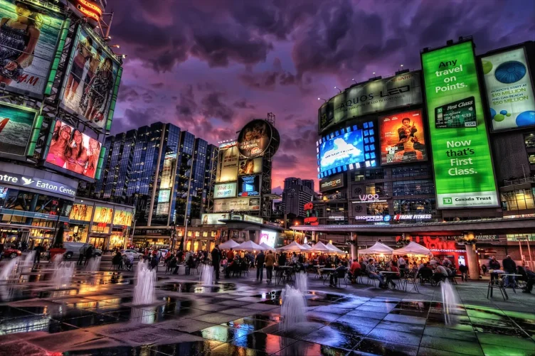 Praça Sankofa Square à noite com letreiros iluminados e fontes no chão.